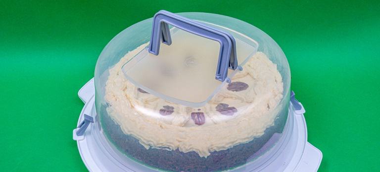 Round transparent plastic cake cover box with gray handles on the top for transportation with a cake inside on a green background