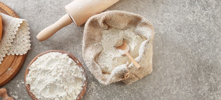 Bowl of wheat flour and sackcloth bag with scoop on light background