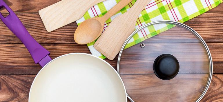 Purple ceramic frying pan with glass lid, kitchen wooden utensil and towel on the wooden table