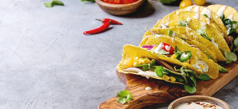 Variety of vegetarian corn tacos with vegetables, green salad, chili pepper served on olive wooden cutting board with tomato and cream sauces with ingredients above over grey texture background.