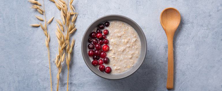 Healthy oats porridge with cranberries on light blue background with oatmeal ears and wooden spoon